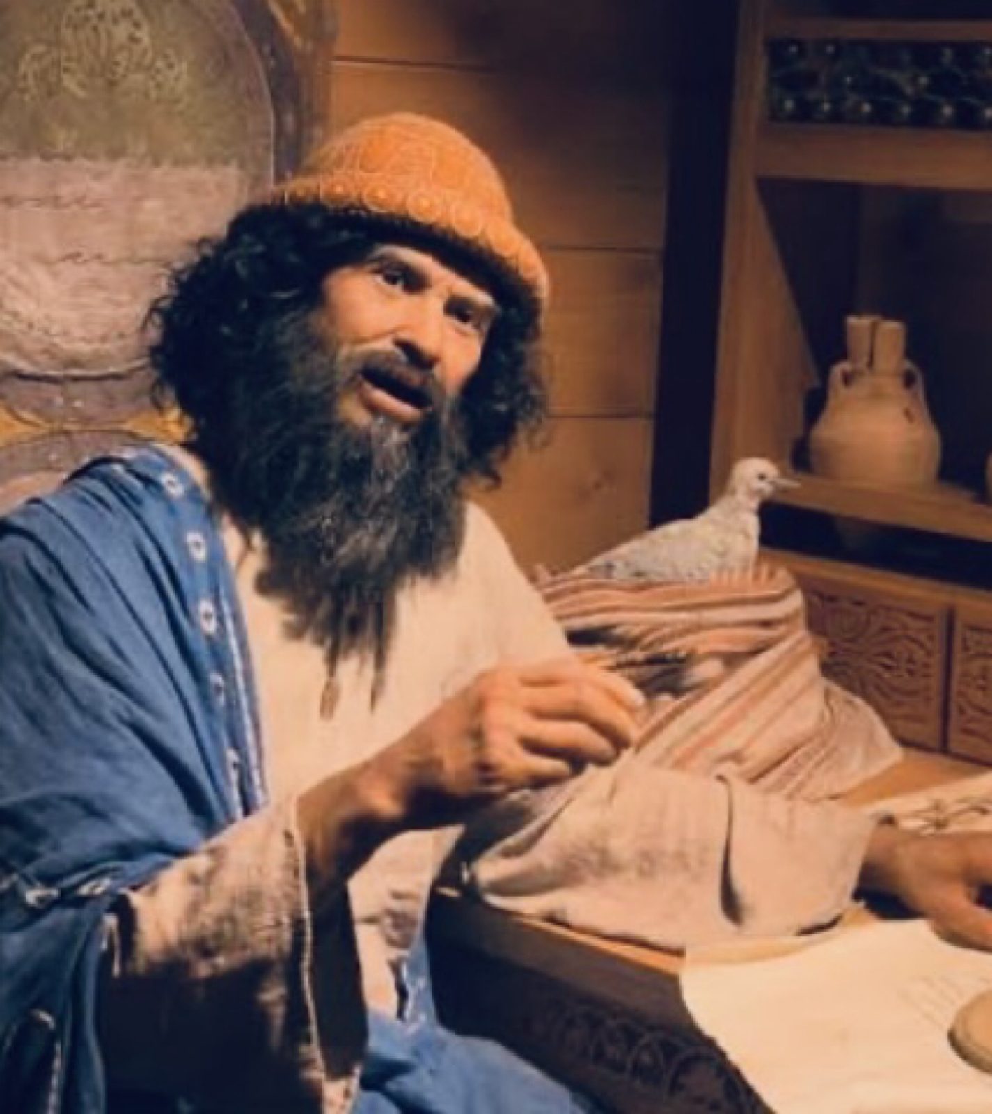 Noah in the orange knit cap looks up from the table, one hand on folded cloth, a white dove beside clay jars.
