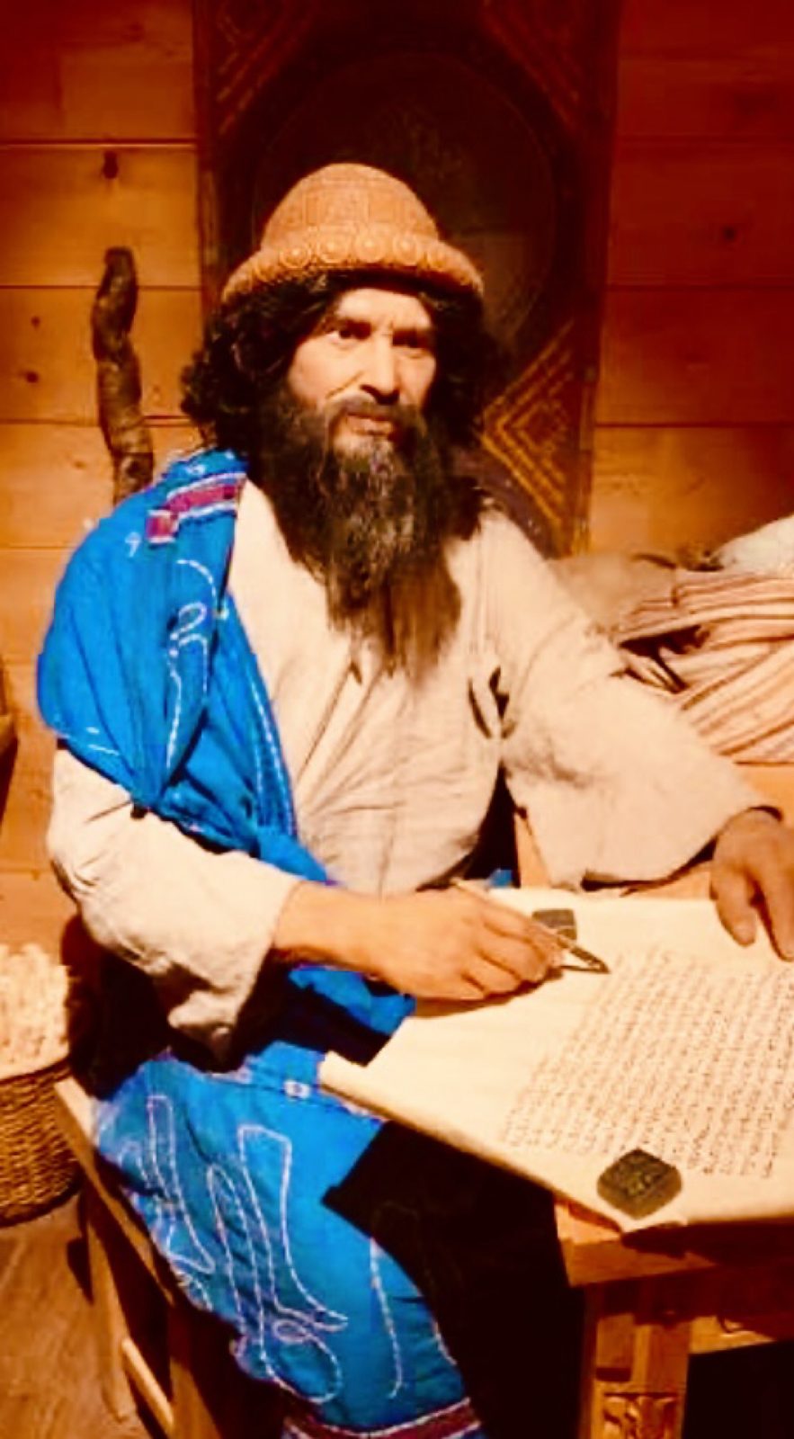 Noah seated with a large written scroll, knit cap and blue robe, stylus in hand.